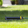 An oscillating lawn sprinkler is watering the grass, casting a spray of water droplets in an arch pattern, connected to a green hose on sunny day.