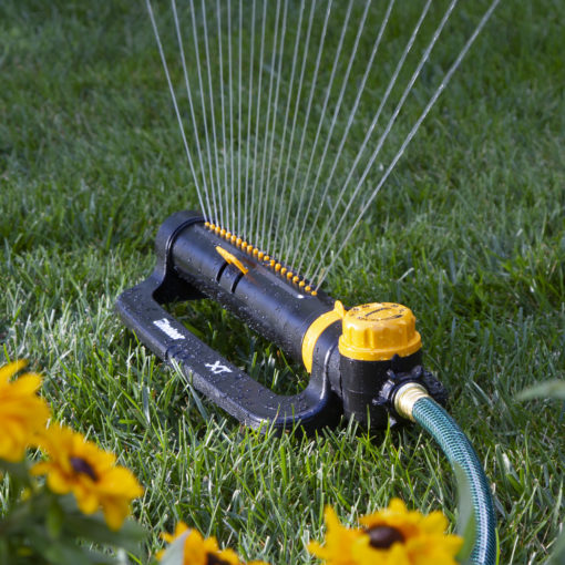 An oscillating lawn sprinkler is actively watering the grass, with a focused spray pattern. Lush green grass and vibrant yellow flowers are at the edges.