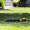 An oscillating sprinkler connected to a green hose waters a lush green lawn with a fine spray of water in a residential garden.