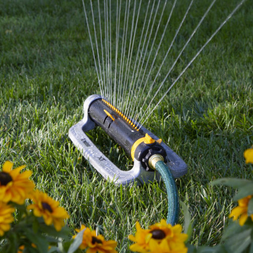 An oscillating sprinkler is watering the grass, surrounded by vibrant yellow and brown flowers, likely marigolds, with water droplets creating a fan-like pattern.