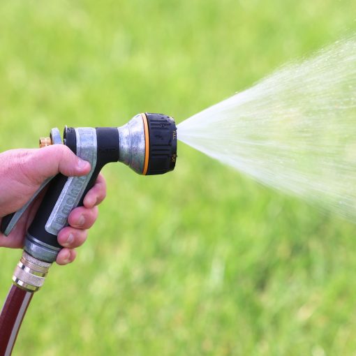 A person's hand is holding a garden hose with a spray nozzle, watering bright green grass, with a fine mist of water visible in the air.