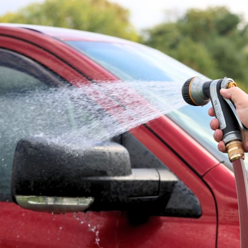 A person's hand holds a hose with a nozzle, spraying water on a red car's window. The vehicle appears to be undergoing outdoor cleaning.