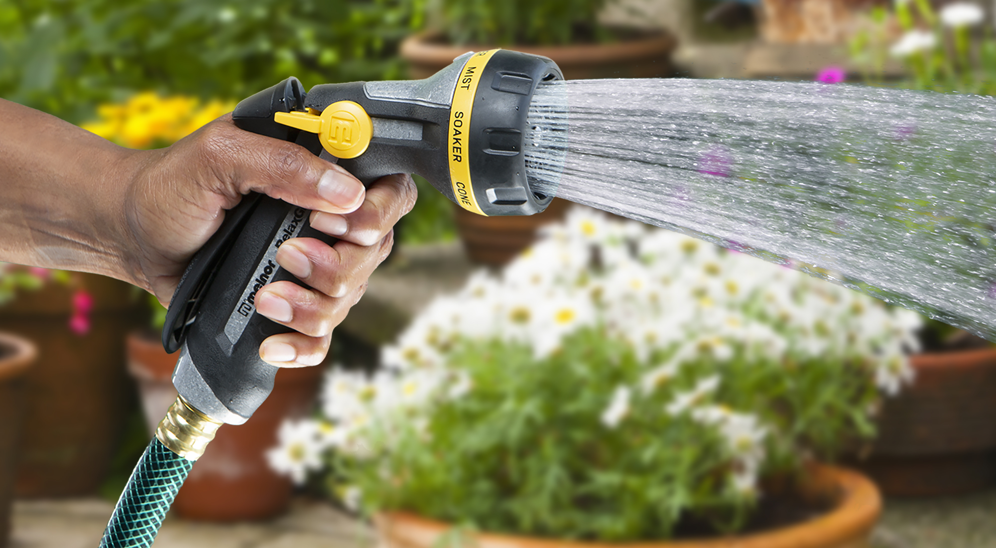 A person is holding a garden hose with a nozzle, spraying water on plants. Behind the stream, potted flowers and greenery indicate a garden setting.