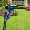 A person's hand holds a blue garden hose nozzle, spraying water onto a lush green lawn, with colorful flowers and a white fence in the background.