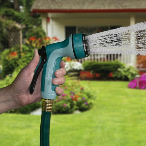 A person is holding a garden hose nozzle, spraying water in a flat pattern. It's a sunny day in a residential yard with a house and flowers.