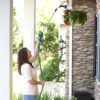 A person is watering hanging red flowers with a telescopic watering wand, standing on a porch by stone pillars and a black lantern.