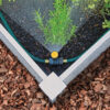 This image shows a garden bed with green plants, covered by a black ground cover fabric. A green garden hose is attached to a brass splitter.