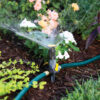 The image depicts a garden sprinkler attached to a green hose, actively watering plants with a fine mist, surrounded by a variety of colorful flowers and greenery.