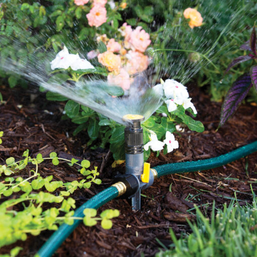 The image depicts a garden sprinkler attached to a green hose, actively watering plants with a fine mist, surrounded by a variety of colorful flowers and greenery.