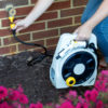 A person is attaching a hose to a spigot outside a brick house, with a portable hose reel nearby and colorful flowers in the foreground.