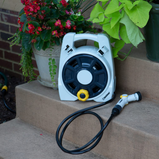 A portable, white and blue hose reel with a yellow and gray nozzle rests on concrete steps beside colorful flowers in plant pots.