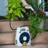 A white portable hose reel sits on wooden steps next to potted plants. A person waters plants using a hose with a spray nozzle.