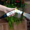 A person's hand is holding a white and yellow garden hose nozzle, spraying water, with plants and a brick wall softly blurred in the background.