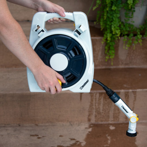 A person's hands hold a white and black automatic hose reel with a nozzle attached; it's being used to water a concrete surface near green plants.