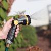 A person's hand is holding a garden hose with a nozzle attachment, spraying water, likely watering plants, in an outdoor setting with greenery.