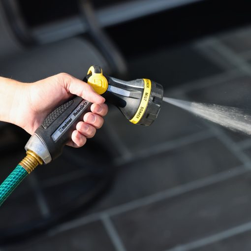 A person's hand is holding a black and yellow garden hose nozzle, spraying water onto a dark tiled surface in daylight.