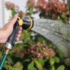 A person is holding a garden hose with a spray nozzle, watering blooming plants. Sunlight creates a bright, fresh outdoor gardening scene.