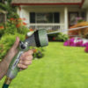 A person's hand is holding a garden hose with a spray nozzle, watering the lawn in front of a house with a porch and colorful flowerbeds.