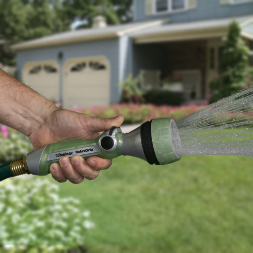 A person's hand is holding a garden hose with a nozzle, spraying water onto grass in front of a suburban house with a double garage door.