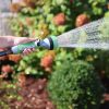 A person's hand holding a garden hose with a spray nozzle, watering plants on a sunny day. Bright sunlight enhances the water droplets' sparkle.