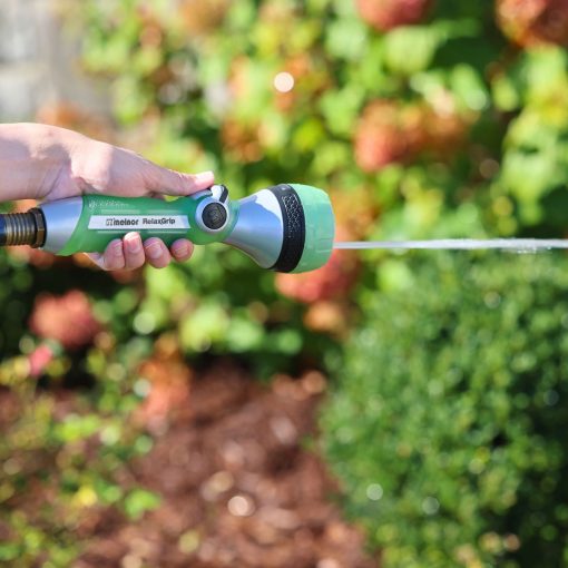 A person is holding a green and black garden hose nozzle, spraying a jet of water towards some lush plants in sunny weather.