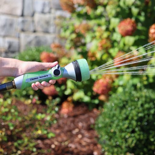 A person's hand is holding a green and gray garden hose nozzle, spraying water in a fan pattern, with foliage and a stone wall in the background.