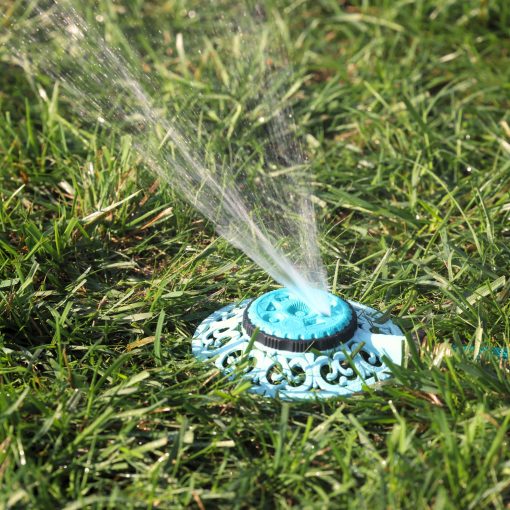 A close-up of a blue lawn sprinkler in operation, with streams of water spraying out, set amidst lush green grass highlighted by sunlight.