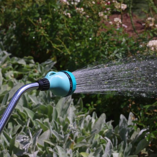 A garden hose with a blue nozzle is spraying water over green plants in a sunny garden with blooming flowers in the background.