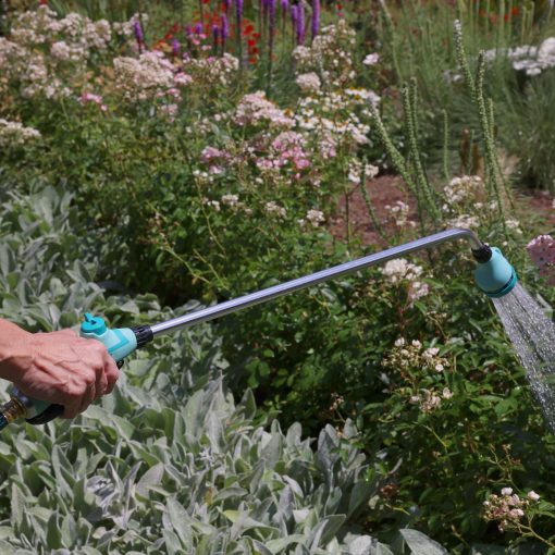 A person's hand is holding a garden hose with a spray attachment, watering a vibrant garden full of various flowers and plants on a sunny day.