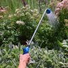 A person is holding a blue watering wand, which is spraying water onto a vibrant garden full of various flowers and plants on a sunny day.