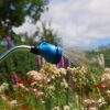 A garden hose with a blue nozzle spraying water in a misting pattern over colorful flowers under a sunny sky with fluffy white clouds.