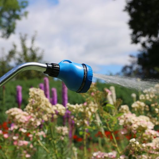 A blue garden hose nozzle is spraying water amidst a sunny garden with pink and purple flowers blooming. Clouds are visible in the blue sky.