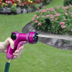 A person is holding a bright pink garden hose nozzle, spraying water onto a lush green lawn surrounded by vibrant pink and red flowers.