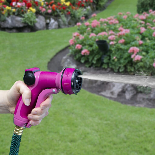 A person is holding a bright pink garden hose nozzle, spraying water onto a lush green lawn surrounded by vibrant pink and red flowers.