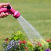 A person is watering colorful flowers using a pink garden hose nozzle. Water is visibly spraying onto the plants against a green grass background.