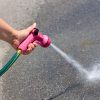 A hand is holding a pink garden hose nozzle, spraying water forcefully onto a sunlit concrete surface, creating a scattered splash pattern.