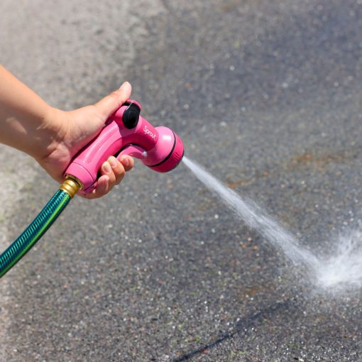 A hand is holding a pink garden hose nozzle, spraying water forcefully onto a sunlit concrete surface, creating a scattered splash pattern.