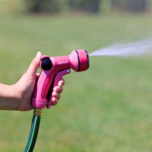 A person is holding a pink garden hose nozzle, spraying water onto a grassy field with a clear sky in the background.