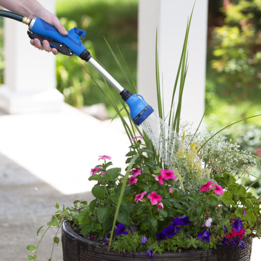 A person's hand is holding a blue garden hose with a nozzle, watering a vibrant basket of pink, purple, and white flowers and greenery.