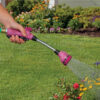 A person is holding a purple garden hose nozzle, spraying water over colorful flowers in a sunlit backyard. Green grass and foliage are visible.