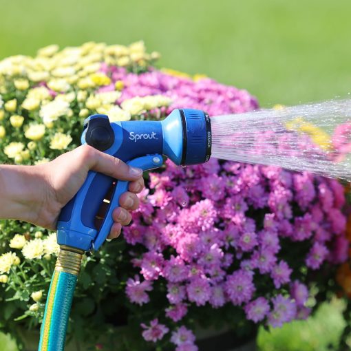 A person's hand is holding a blue garden hose nozzle labeled "Sprout", watering vibrant pink flowers under bright sunlight.