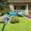 A person's hand is holding a blue and grey garden hose nozzle, spraying water onto a lawn with colorful flowers in front of a house.