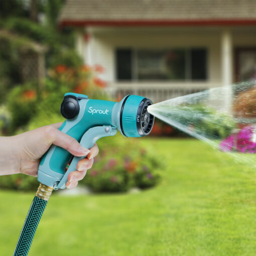 A person's hand is holding a blue and grey garden hose nozzle, spraying water onto a lawn with colorful flowers in front of a house.