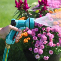 A person's hand is holding a teal garden hose nozzle, labeled "Sprout," spraying water onto blooming pink and orange flowers under sunlight.