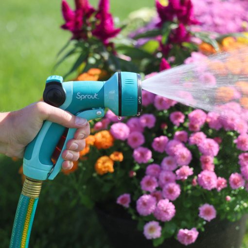 A person's hand is holding a teal garden hose nozzle, labeled "Sprout," spraying water onto blooming pink and orange flowers under sunlight.