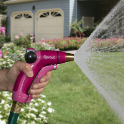 A person's hand holds a pink garden hose nozzle, labeled "Sprout," spraying water towards a lush green lawn in front of a house with garage doors.