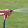 A person is holding a pink garden hose nozzle, spraying water onto a lush green lawn on a sunny day. The focus is on the hand and nozzle.