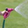 A person is holding a pink garden hose nozzle spraying water onto a lush green lawn with a fine, dispersed mist setting, indicating garden maintenance.