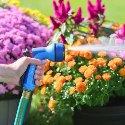 A person's hand is holding a blue garden hose with a spray nozzle, watering vibrant orange flowers backdropped by purple flowers and greenery.