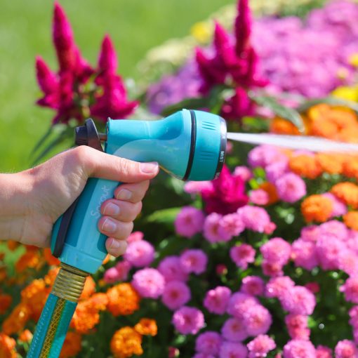 A person's hand is holding a blue garden hose with a spray nozzle, watering vibrant pink and orange flowers in a sunny garden setting.